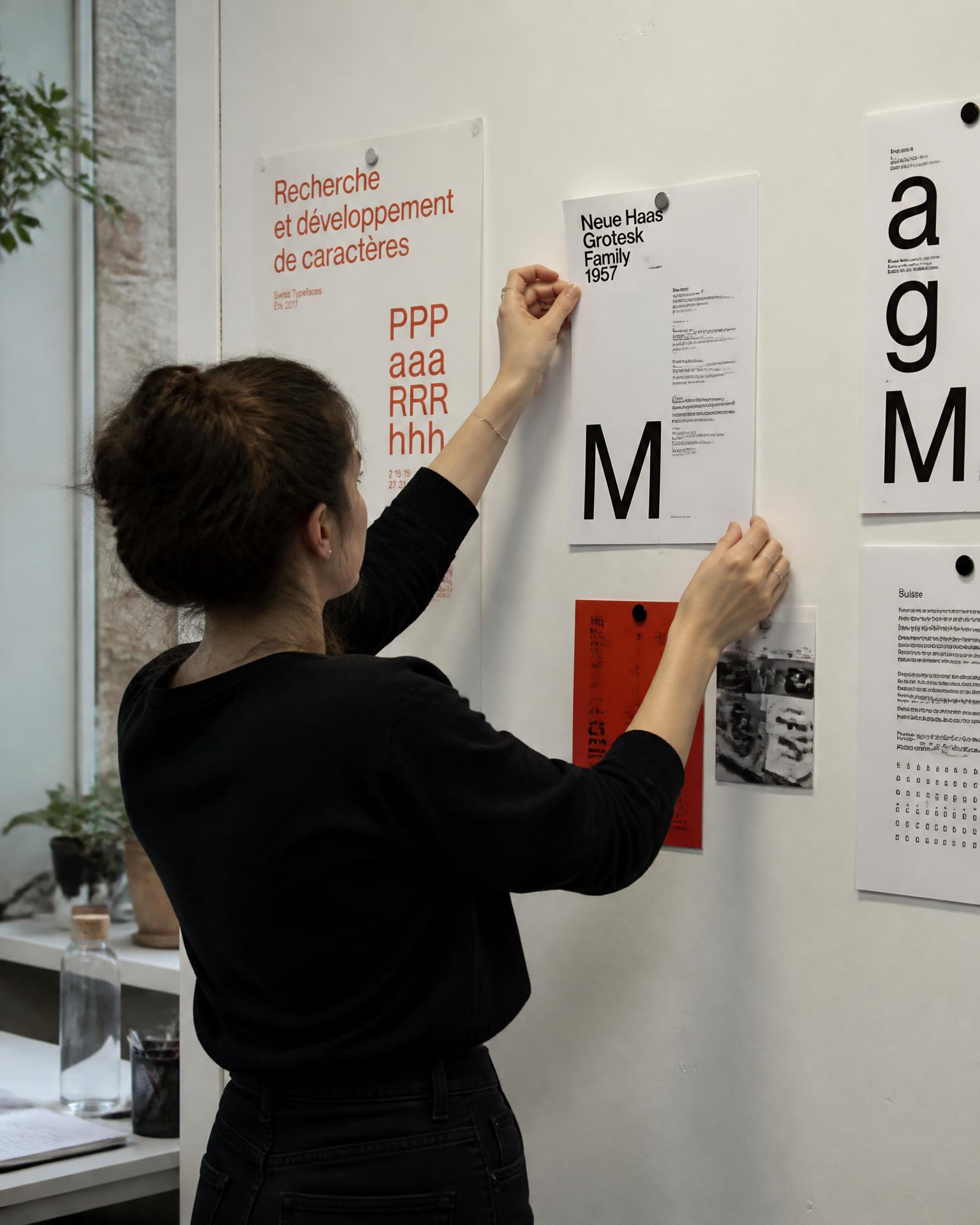 A designer pinning printed type proofs to the studio wall, comparing them against earlier tests